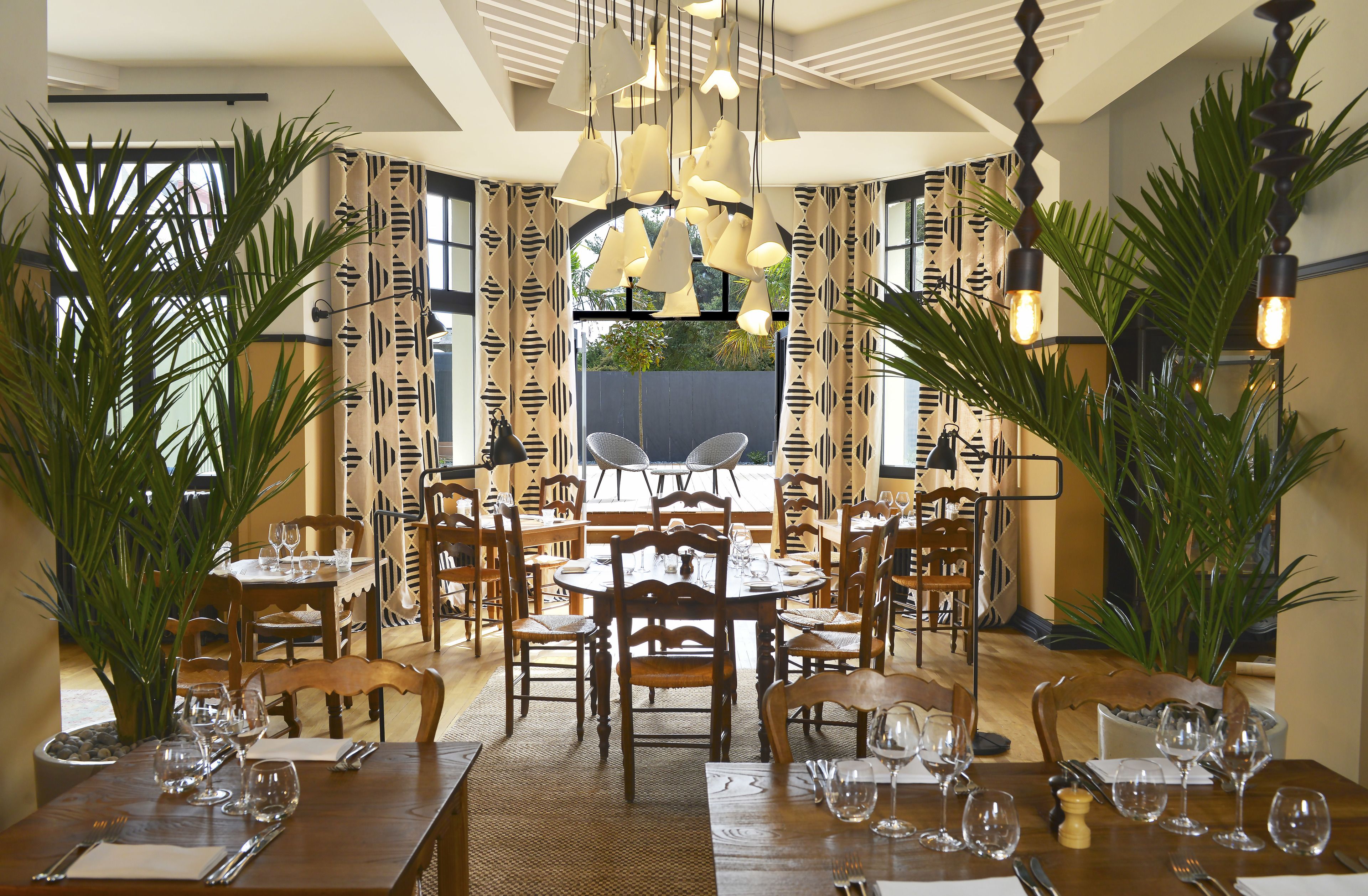 A dining area with multiple tables set for dining, wooden chairs, hanging pendant lights, and large potted plants.