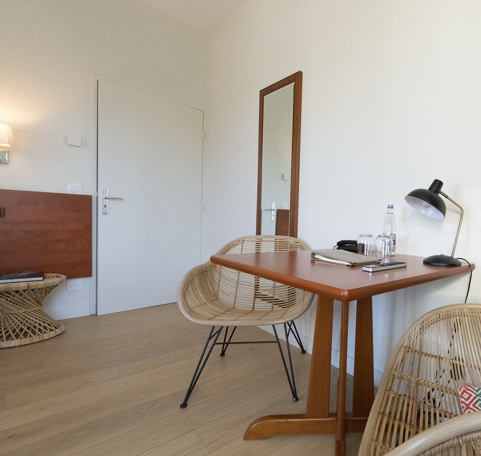 Una habitación pequeña y ordenada con un escritorio de madera, dos sillas de mimbre, un espejo, una lámpara y una mesita con un libro.