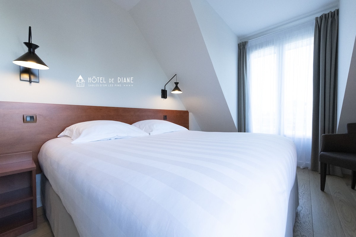 Habitación de hotel con una cama grande, dos lámparas fijadas a la pared, un cabecero de madera, una silla y una ventana con cortinas.