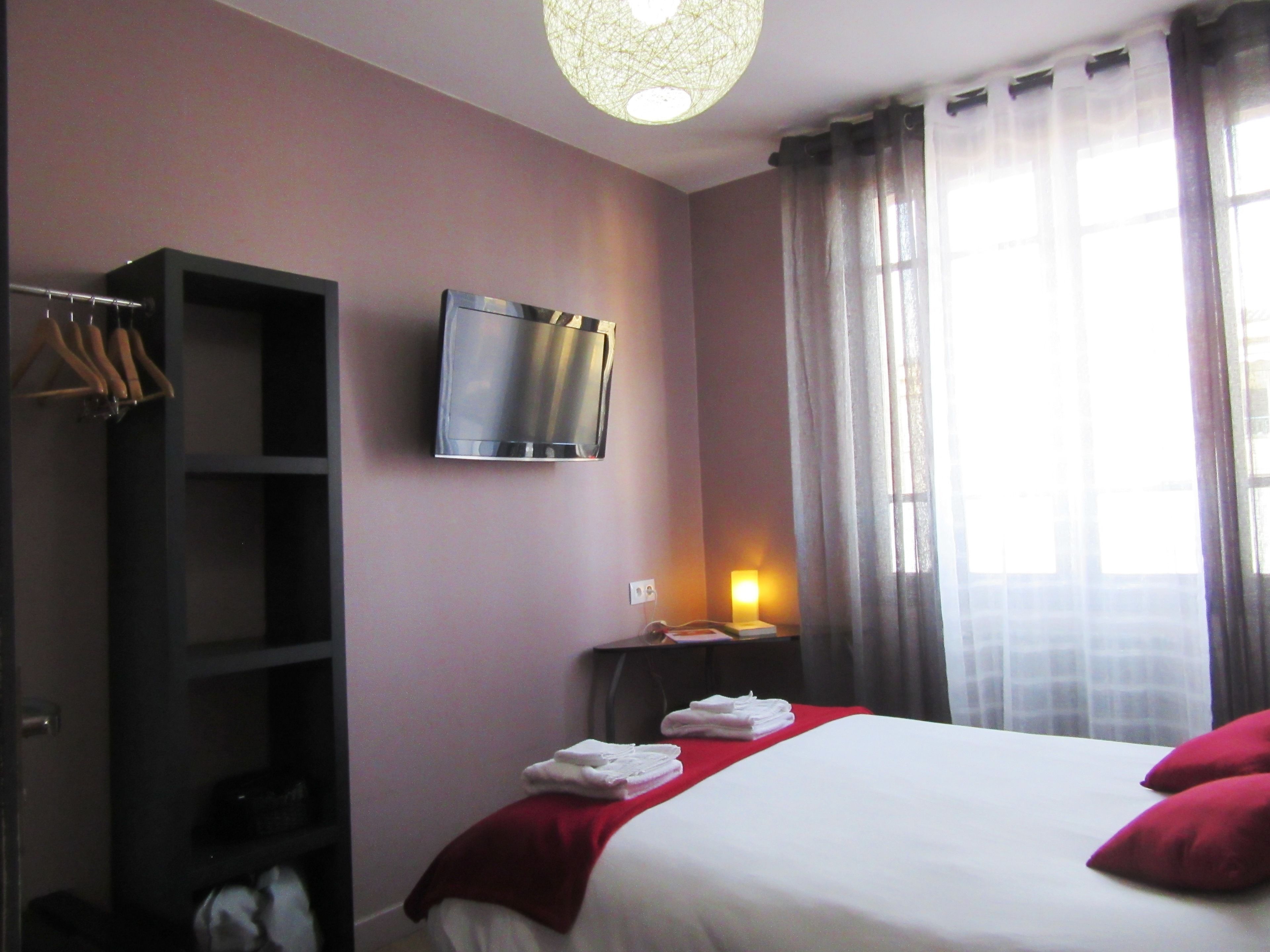 Habitación de hotel con cama, televisor montado en la pared, estantería y ventana con cortinas.