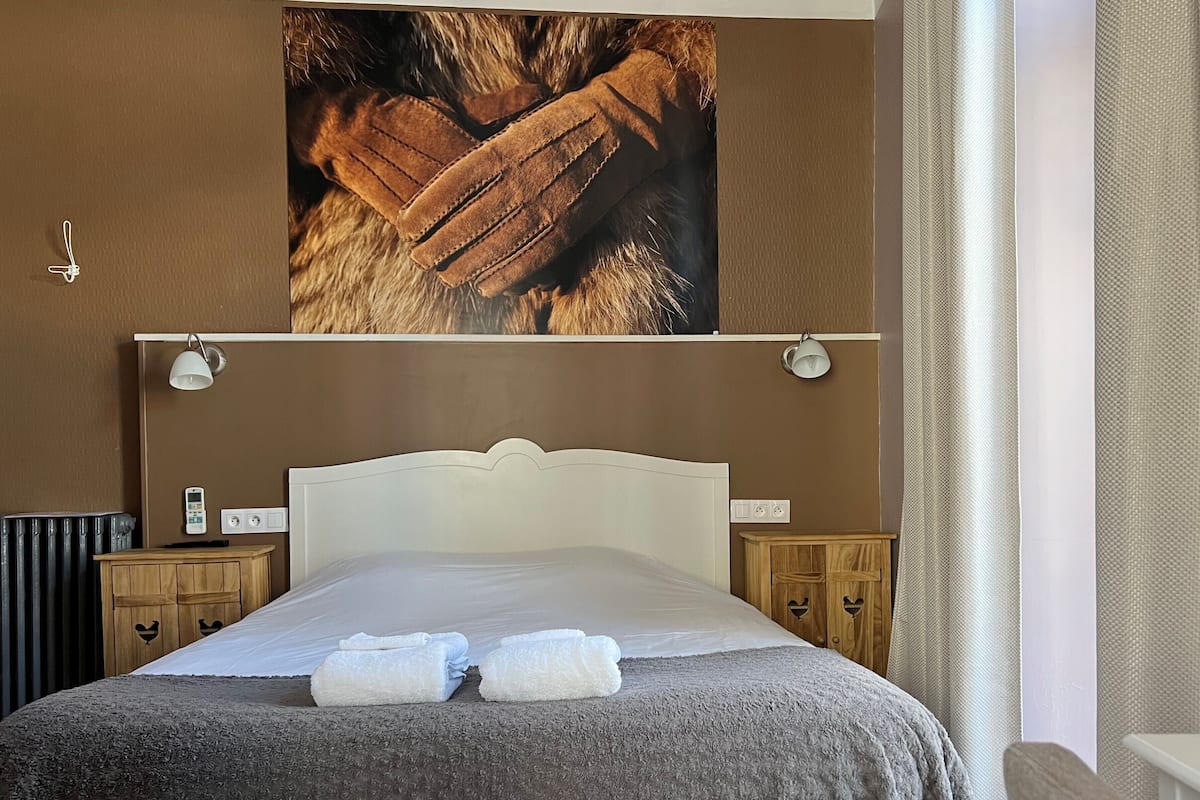 A neatly made bed with a grey blanket and two folded towels, wooden nightstands with lamps, and a large framed picture above the bed.