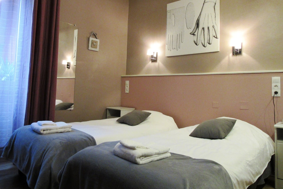 A hotel room with two beds, each with a pillow and a folded towel. There is a wall-mounted light fixture and a painting above the beds.