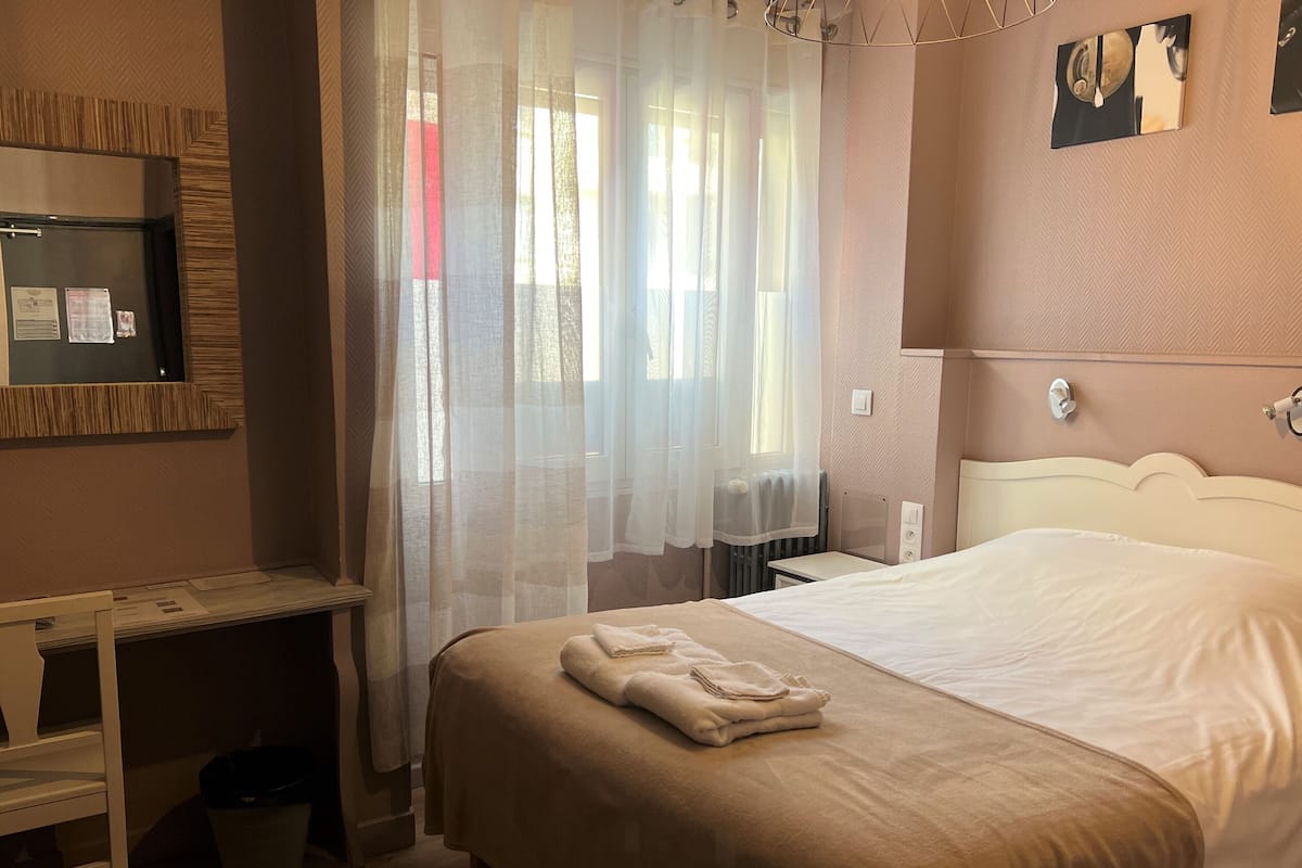 A hotel room with a bed, a bedside table, a mirror, and a window with sheer curtains.