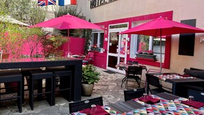 An outdoor seating area with tables and chairs under pink umbrellas, a potted plant, and a sign for Hotel des Causses.