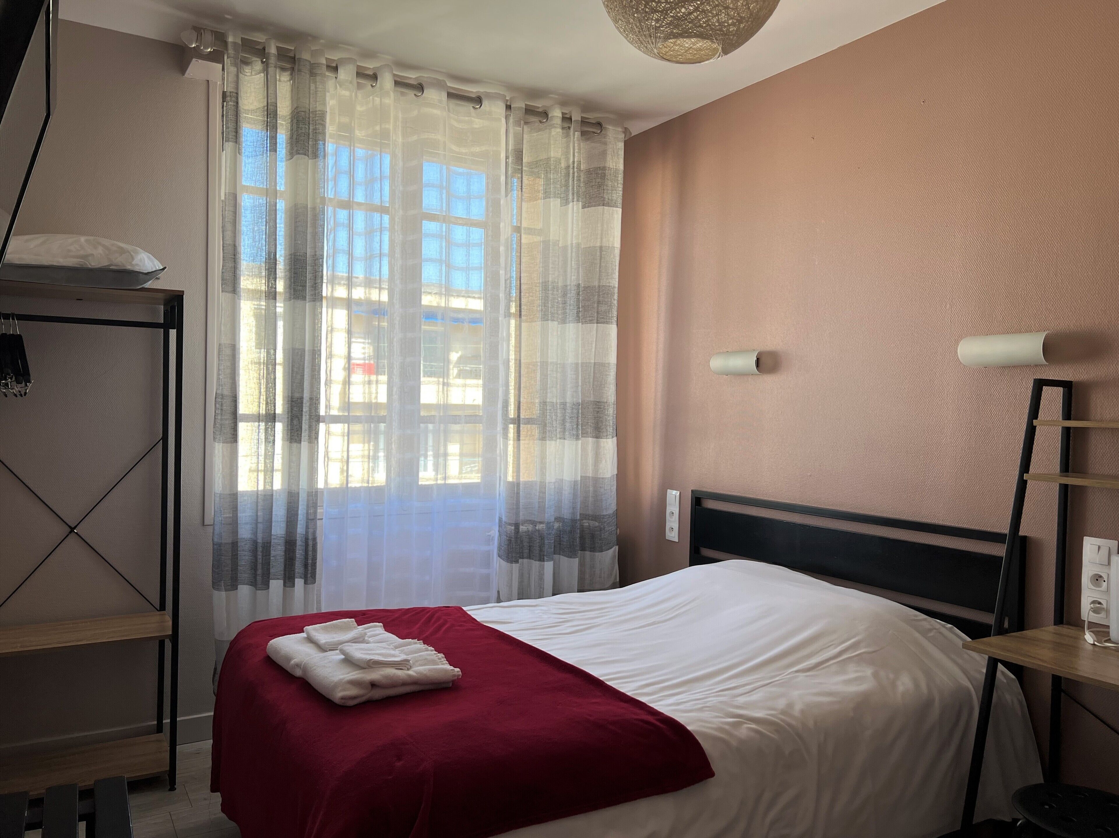 A hotel room with a bed, a red throw blanket, a bedside table, a lamp, and a window with sheer curtains.