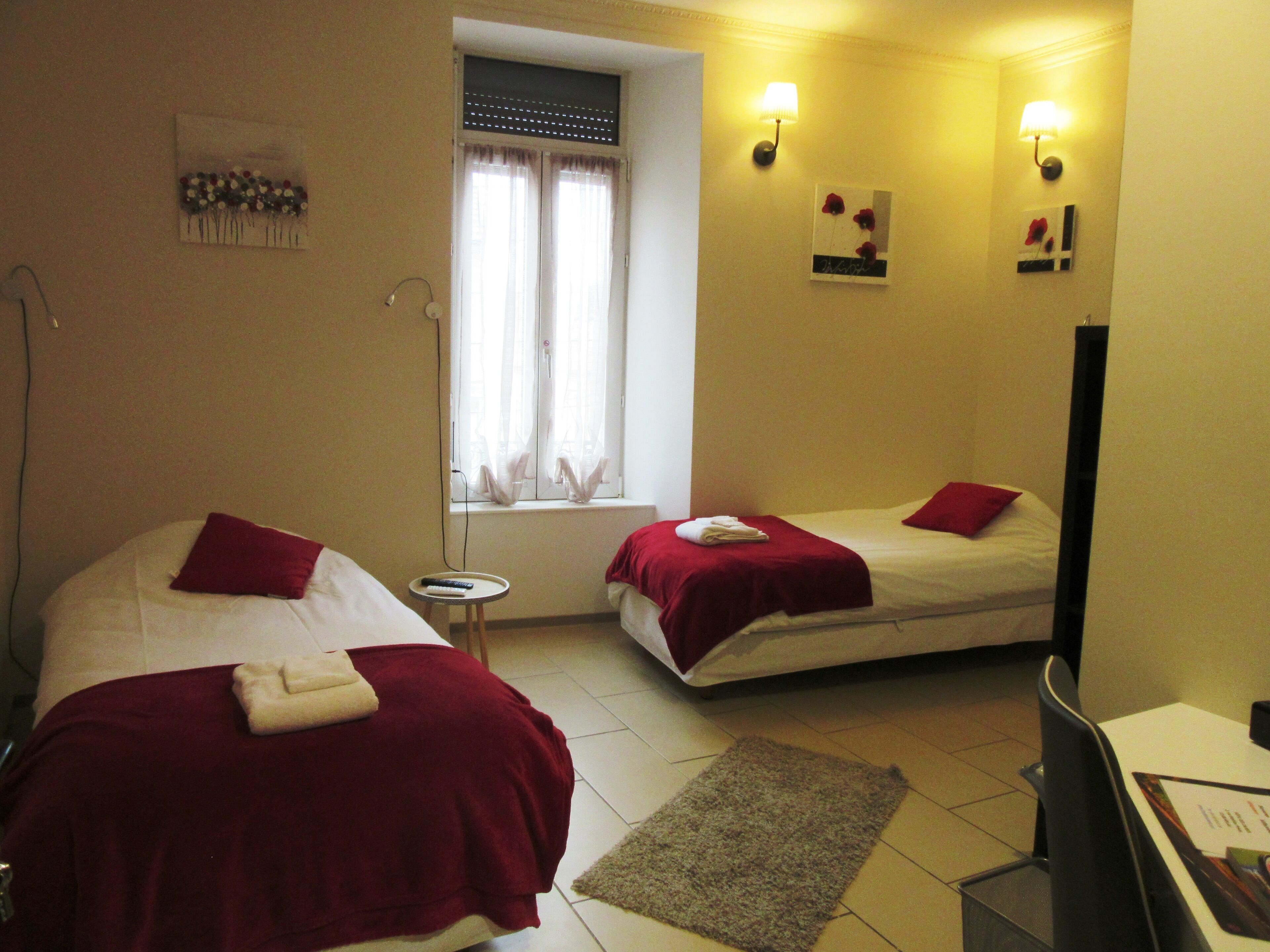 Habitación de hotel con dos camas, cada una con una colcha roja, una mesita redonda con un control remoto y una ventana con cortinas.