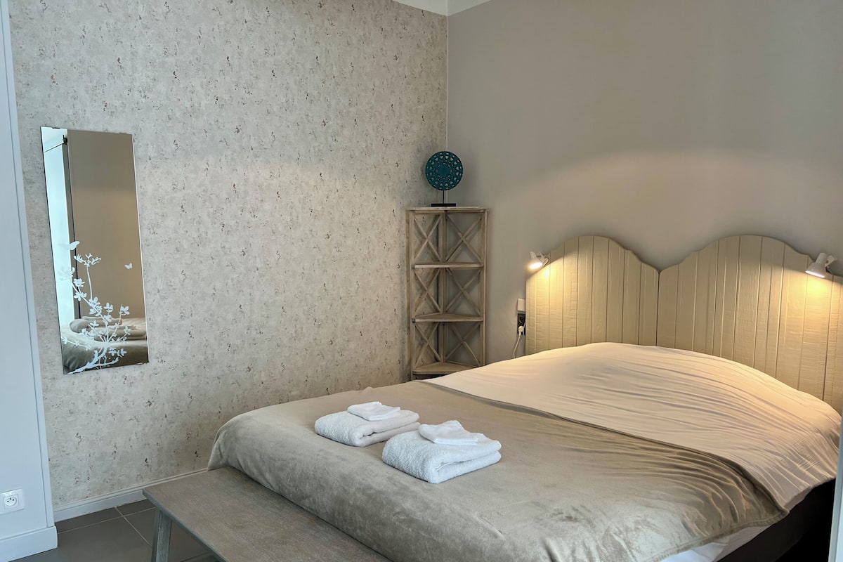 A neatly arranged bedroom with a bed, bench, bedside table, wall-mounted mirror, and a decorative item.