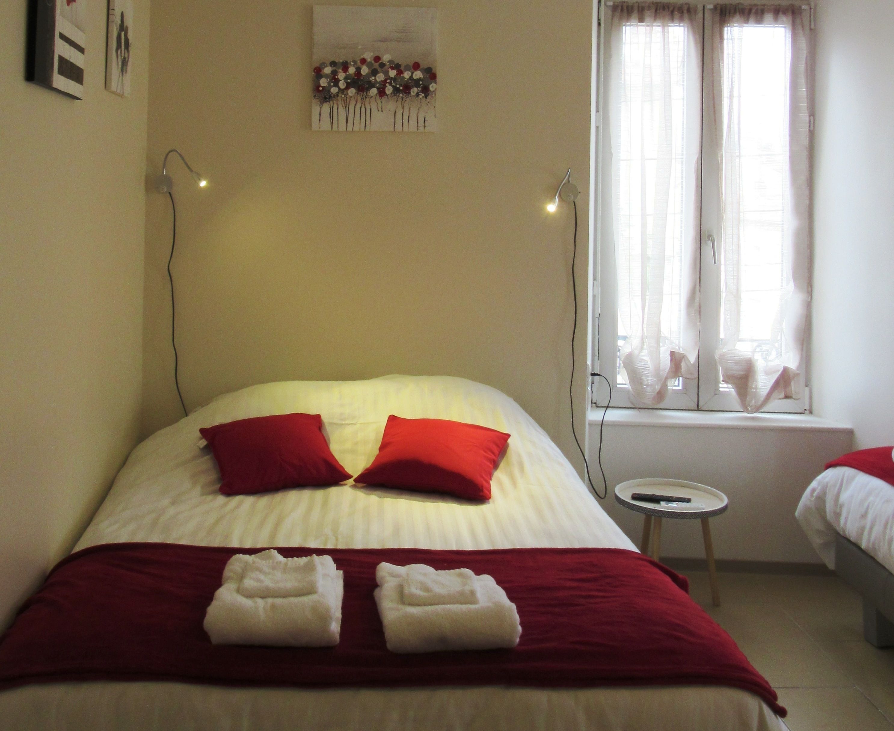 A neatly made bed with red and white bedding, two towels folded at the foot of the bed, a small bedside table with a remote control, and a window with sheer curtains.