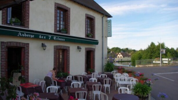 Un espace extérieur aménagé avec des tables et des chaises, un bâtiment sur lequel on peut lire l’inscription « Auberge des 7 Colosés », et une vue sur une rue et la verdure en arrière-plan.