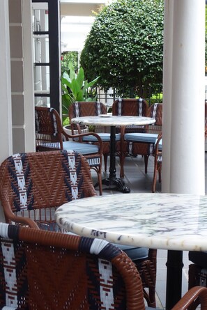 An outdoor seating area with wicker chairs and tables, surrounded by greenery and a white building.