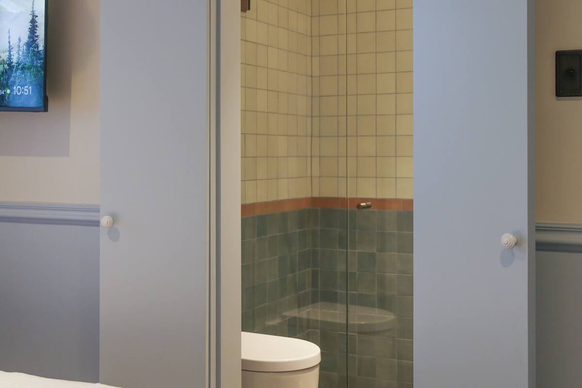Une chambre d’hôtel équipée d’un lit, d’une télévision et d’une douche séparée par une porte en verre.