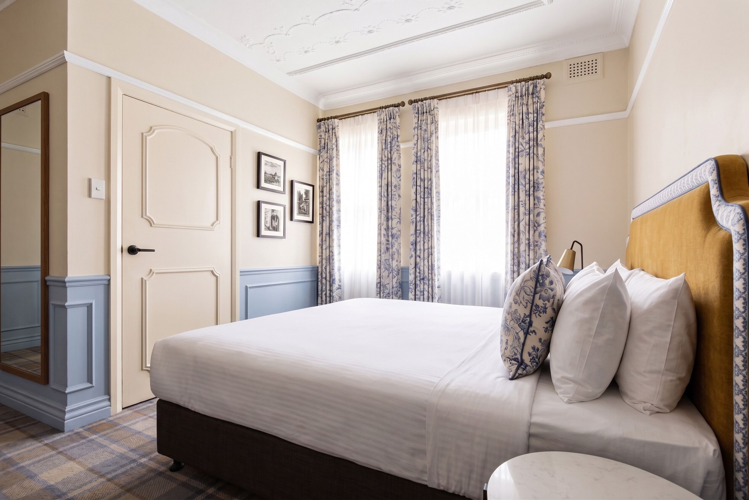A hotel room with a bed, a door, a wall-mounted clock, and a window with curtains.