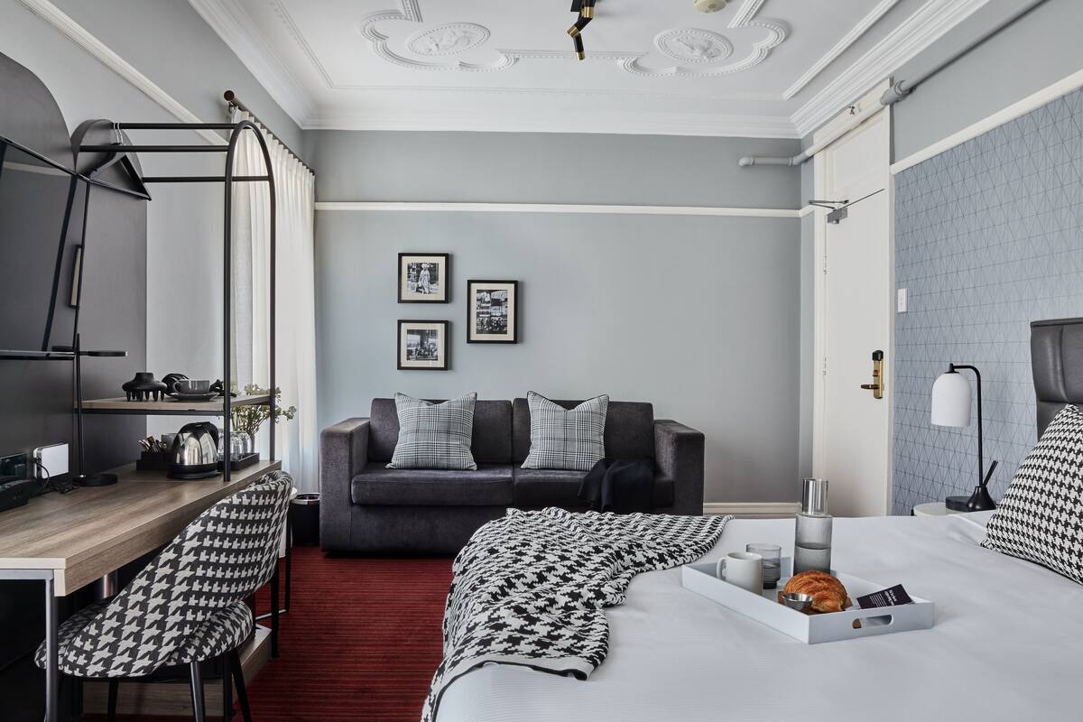 Une chambre d’hôtel moderne avec un canapé gris, un coin repas doté d’un tabouret de bar, un lit avec un plateau de petit-déjeuner et une moquette rouge.