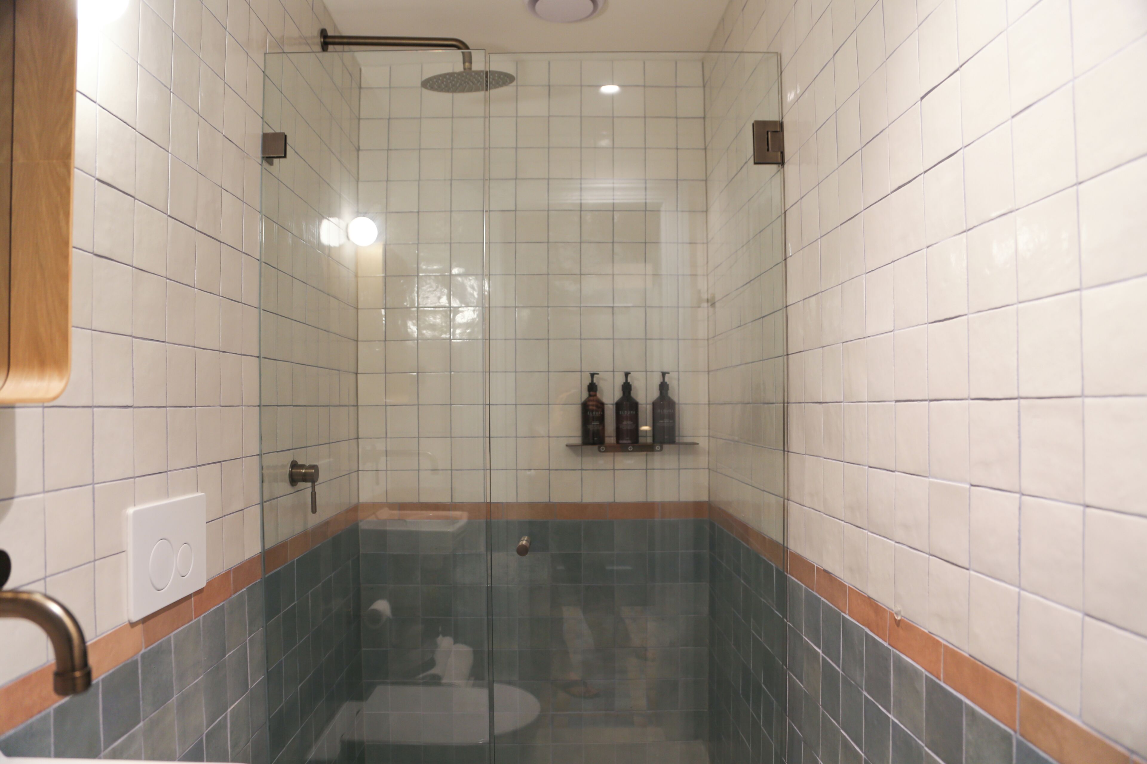 Ein Badezimmer mit weißen Fliesen, einer Duschkabine aus Glas, einem Waschbecken und einem Ablagefach mit Pflegeprodukten.