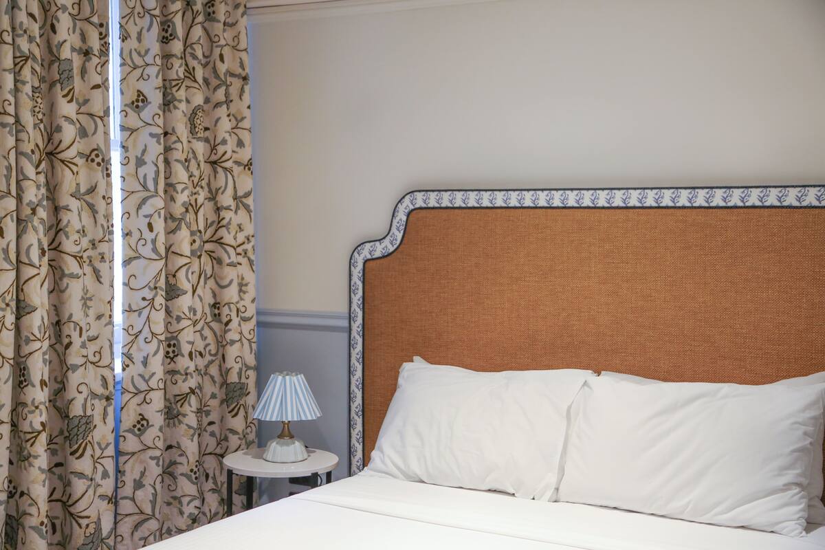 Un lit avec une literie blanche, une tête de lit ornée d’une bordure à motifs et une table de chevet avec une lampe.