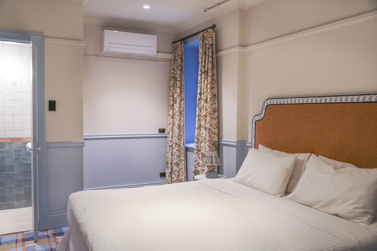 Une chambre d’hôtel avec un lit, des rideaux, un appareil de climatisation et une vue sur l’extérieur, visible à travers une porte.