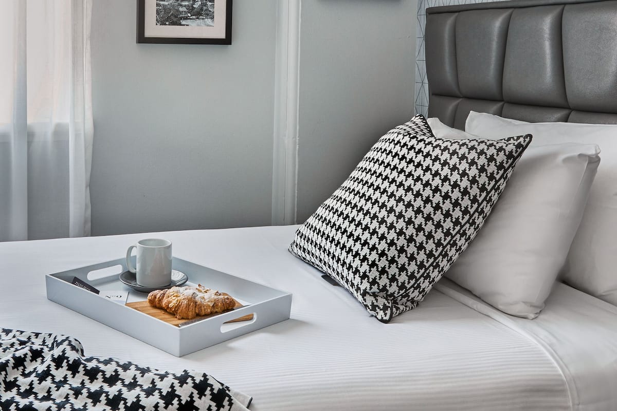 Une chambre d’hôtel bien rangée, avec un lit, un plateau contenant une tasse et une pâtisserie, et deux tableaux encadrés accrochés au mur.