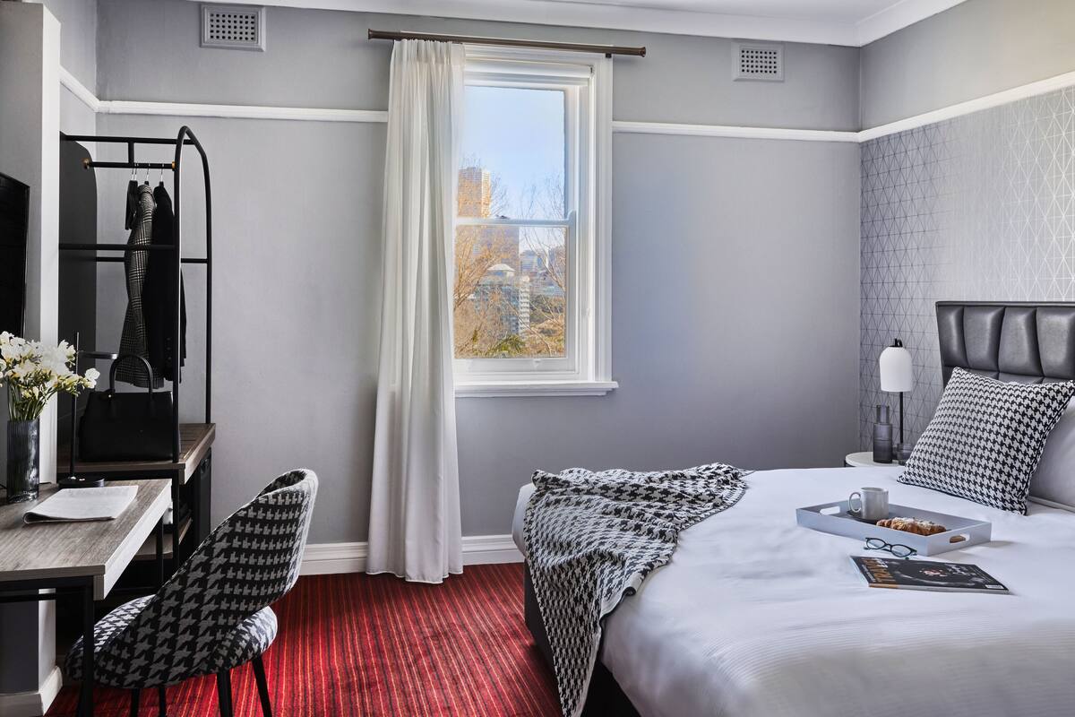 Une chambre d’hôtel avec un lit, un bureau et une chaise, une fenêtre avec des rideaux et une vue sur les bâtiments environnants.