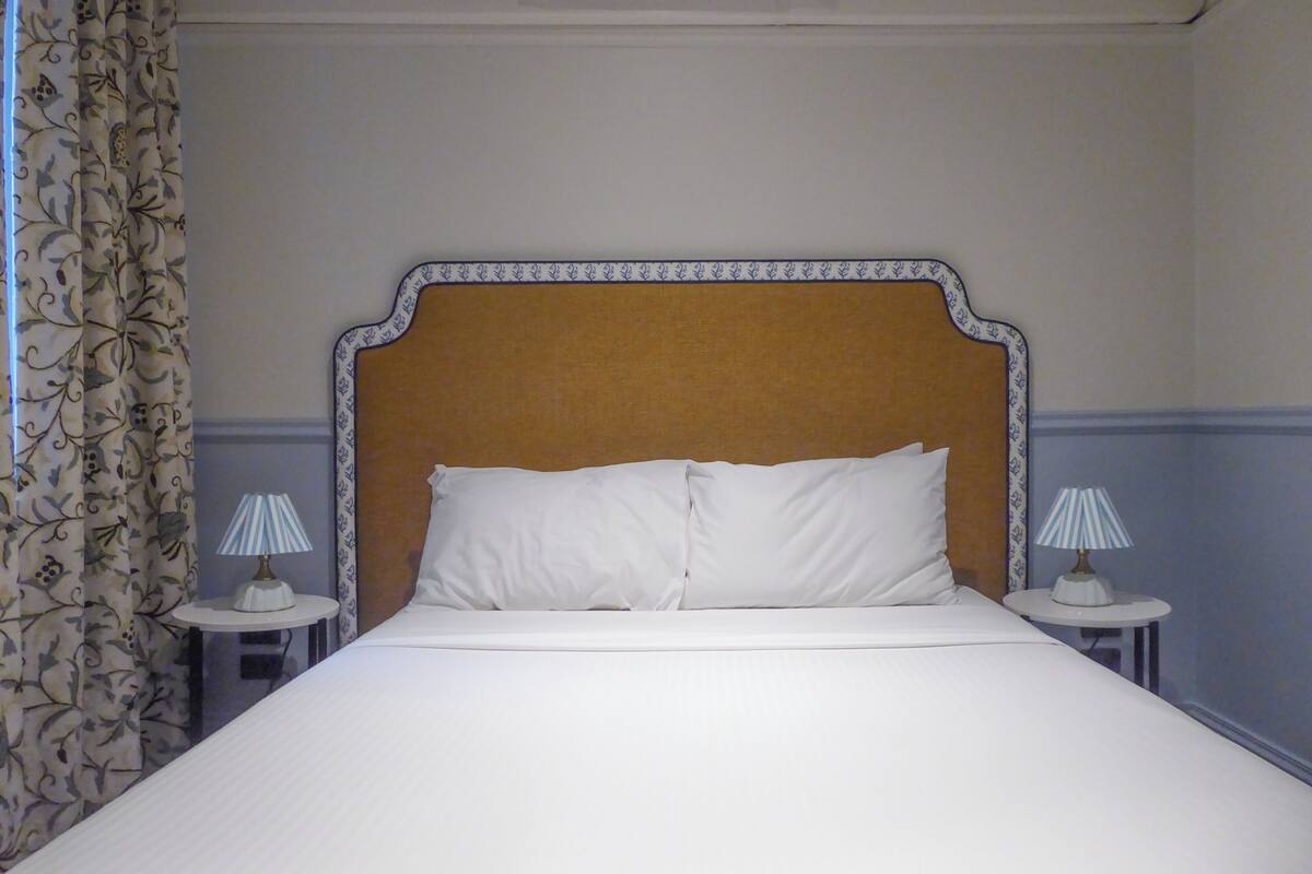 Un lit bien fait, avec une tête de lit, deux tables de chevet dotées de lampes et des rideaux à motifs.