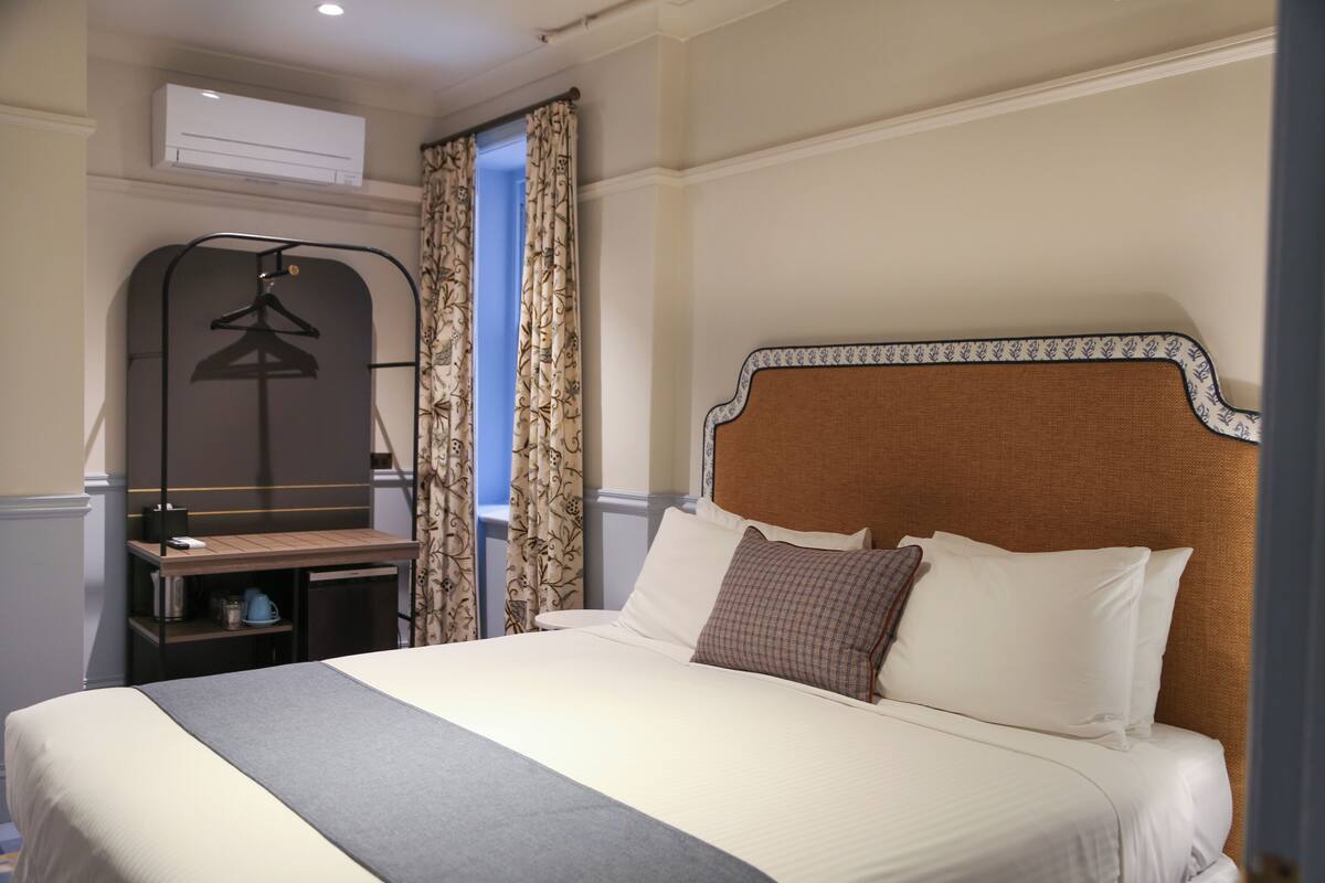 Une chambre d’hôtel comprenant un lit, un bureau avec une chaise, un porte-manteau et une fenêtre avec des rideaux.