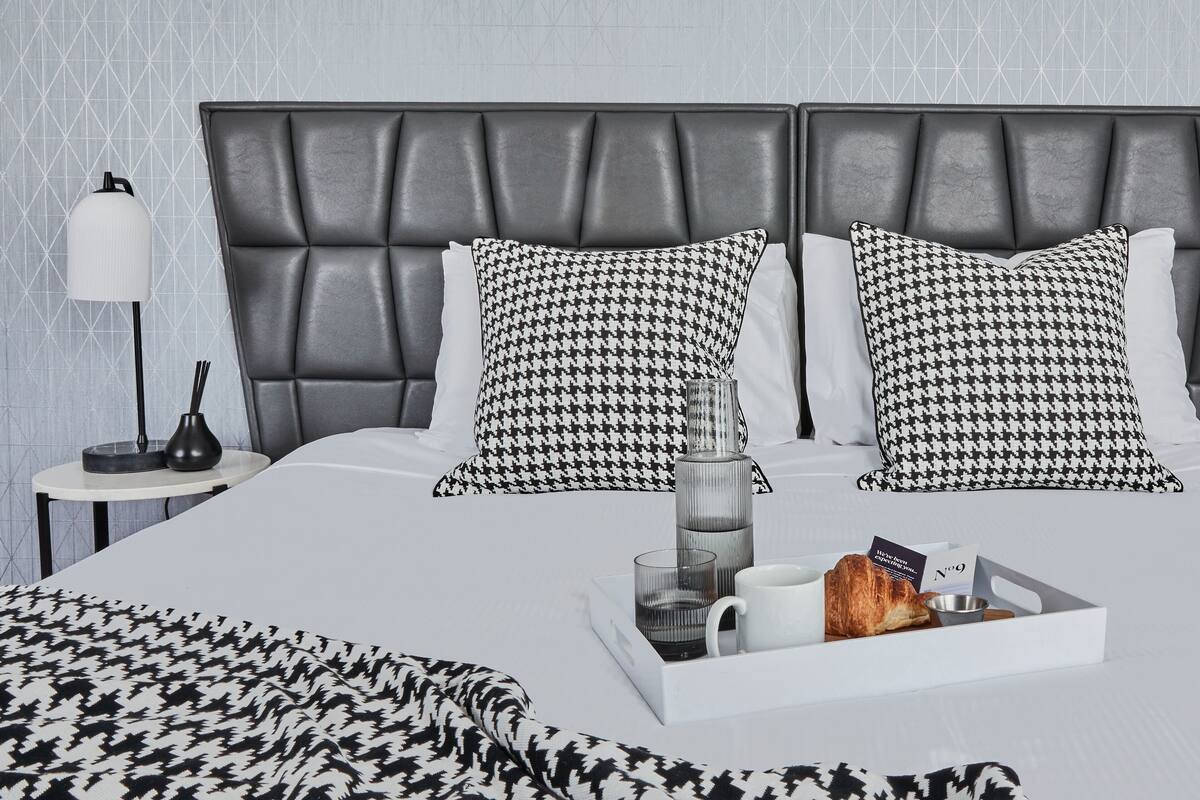 Une chambre d’hôtel moderne avec un grand lit, deux coussins à motif pied-de-poule, une table de chevet avec une lampe et un plateau contenant des articles pour le petit-déjeuner.