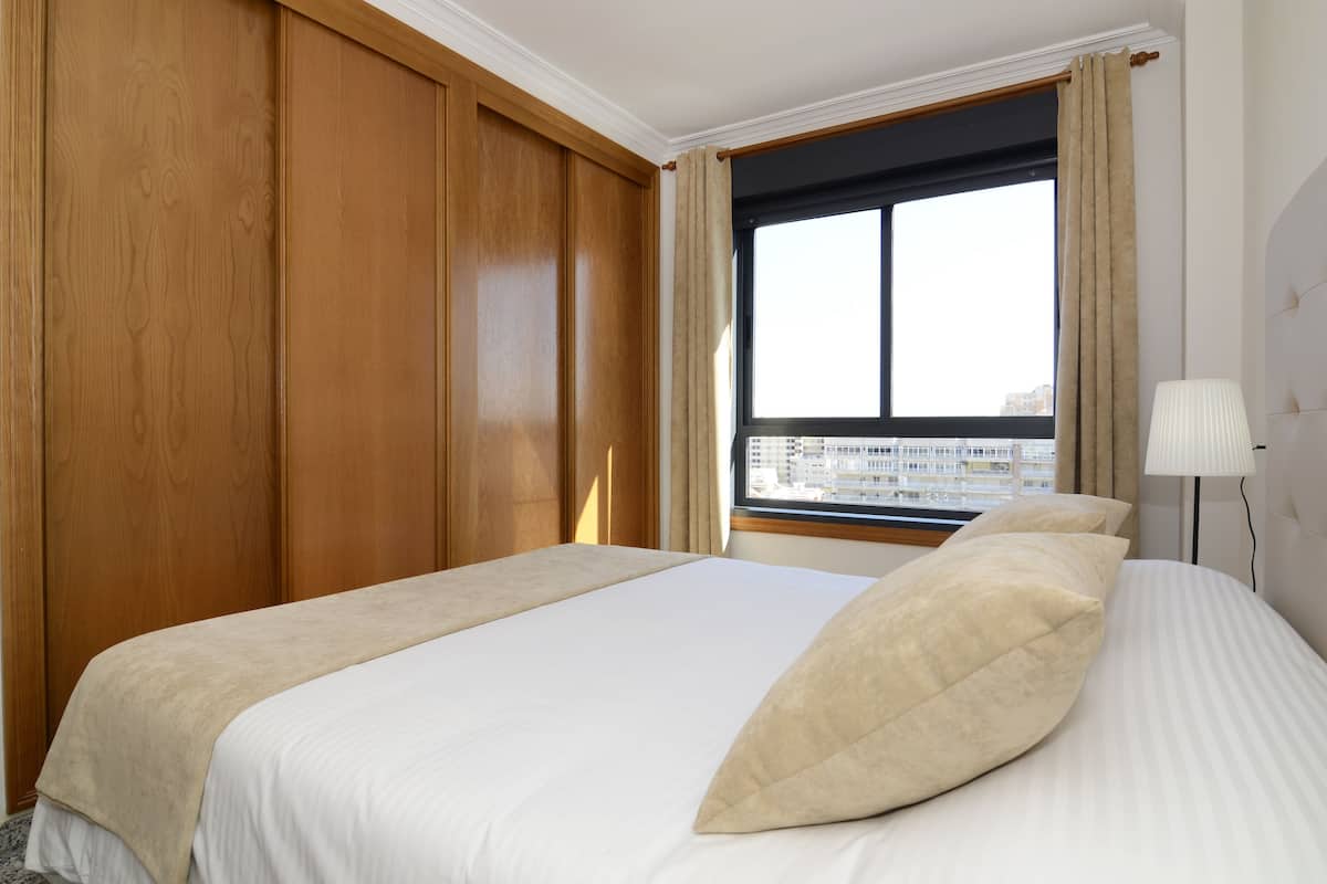 A hotel room with a large bed, wooden paneling, a window with a city view, and a bedside lamp.