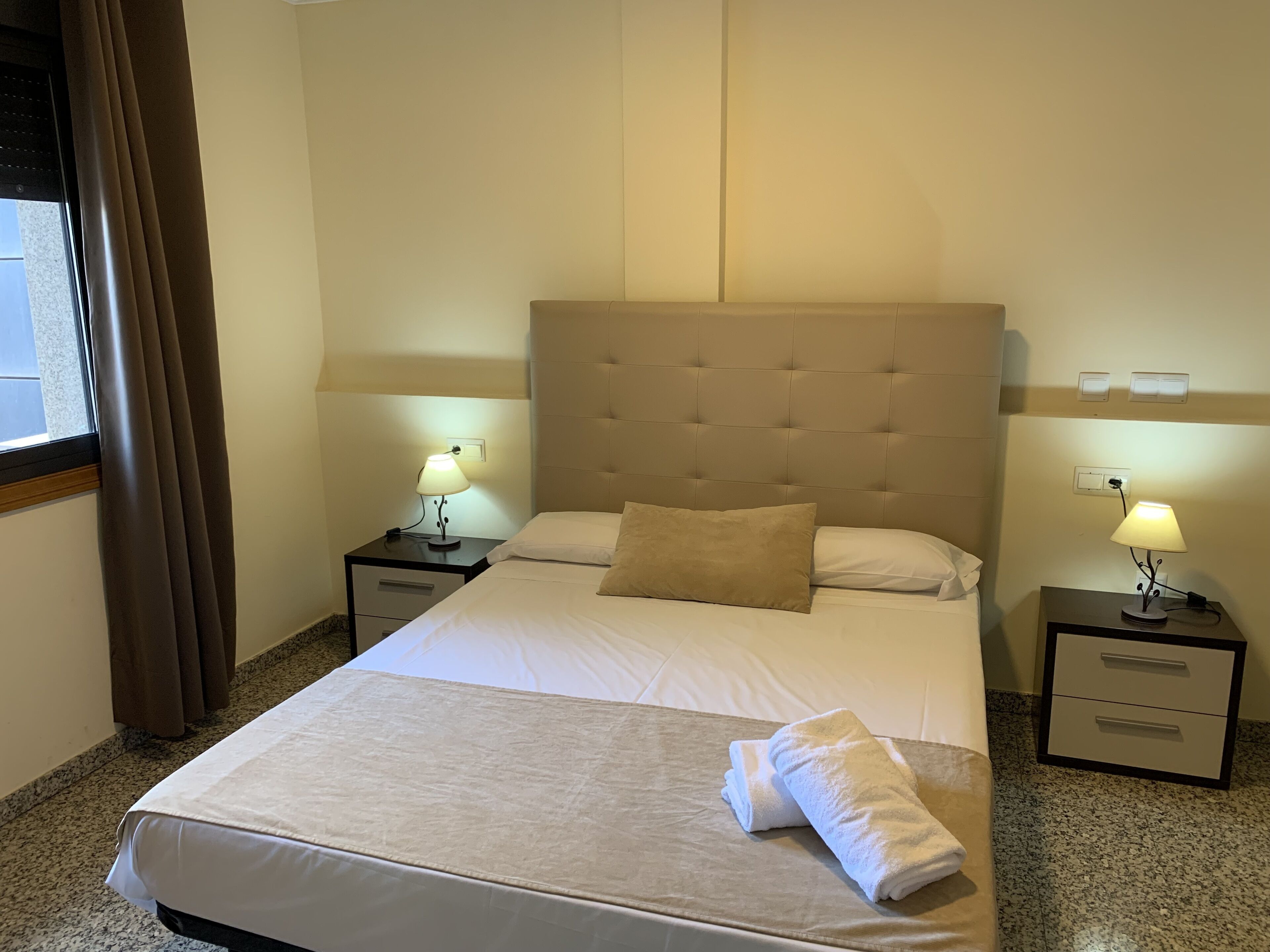 A neatly made bed with a beige headboard, two bedside lamps, a window with curtains, and a small nightstand with a drawer.