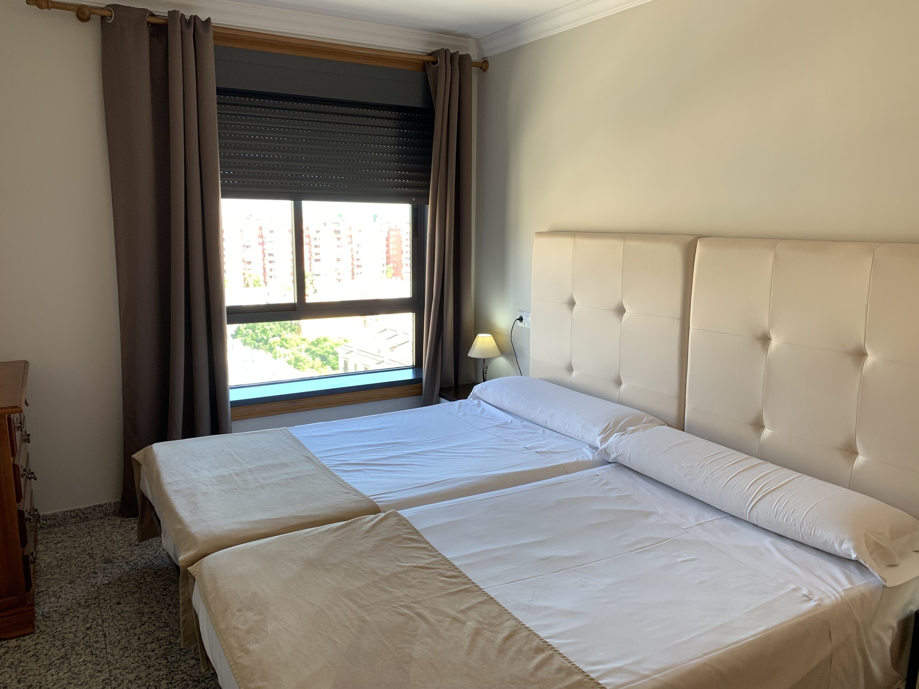 Une chambre d’hôtel avec deux lits, une fenêtre avec des rideaux, une lampe et une vue sur les bâtiments environnants.