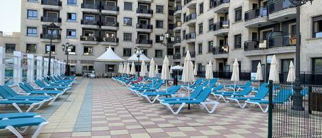 A rooftop pool area with blue lounge chairs and umbrellas, surrounded by a high-rise building with balconies.