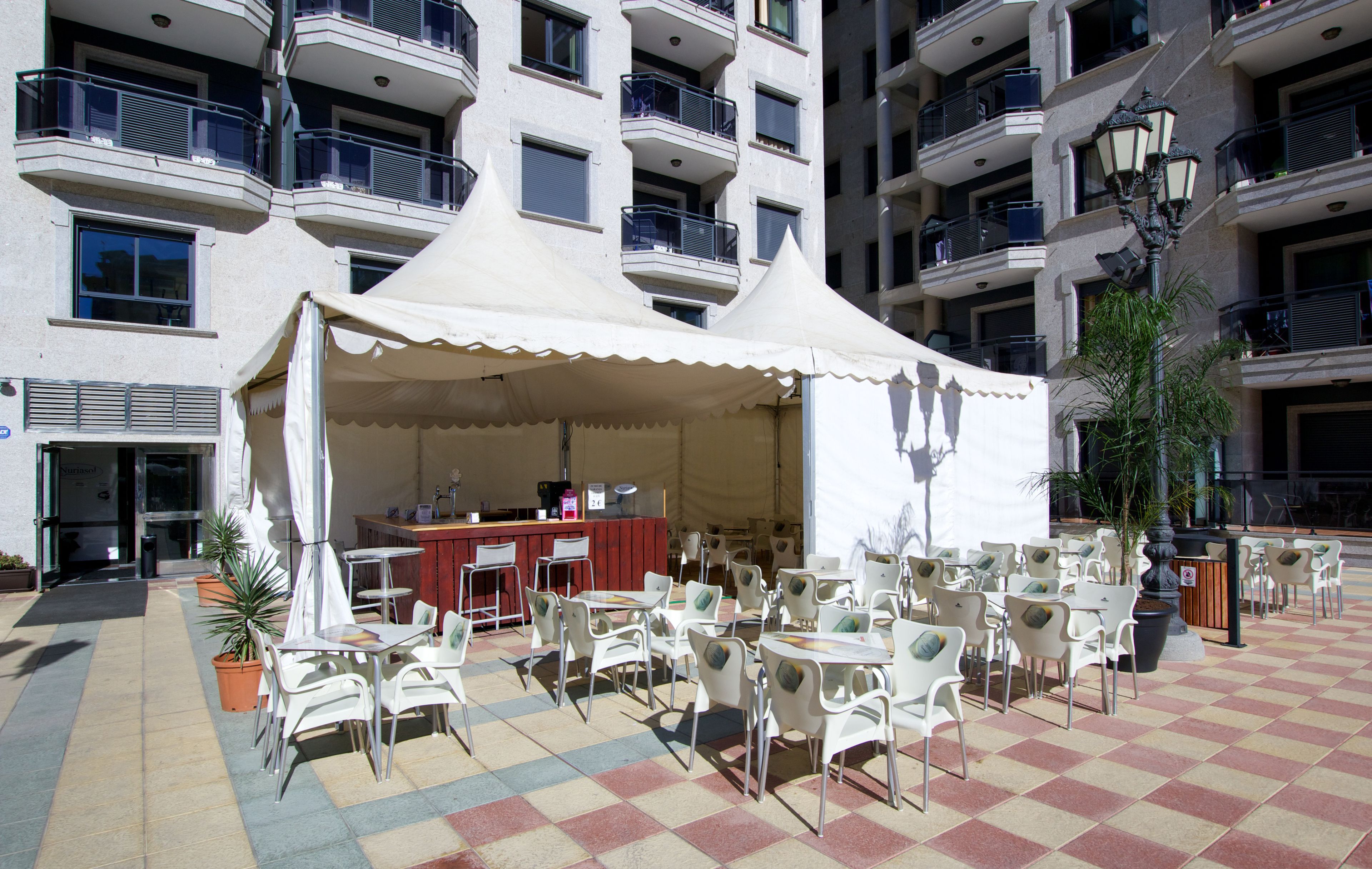 Un café al aire libre con sillas y mesas blancas, una carpa blanca y una zona de bar con mostrador rojo.