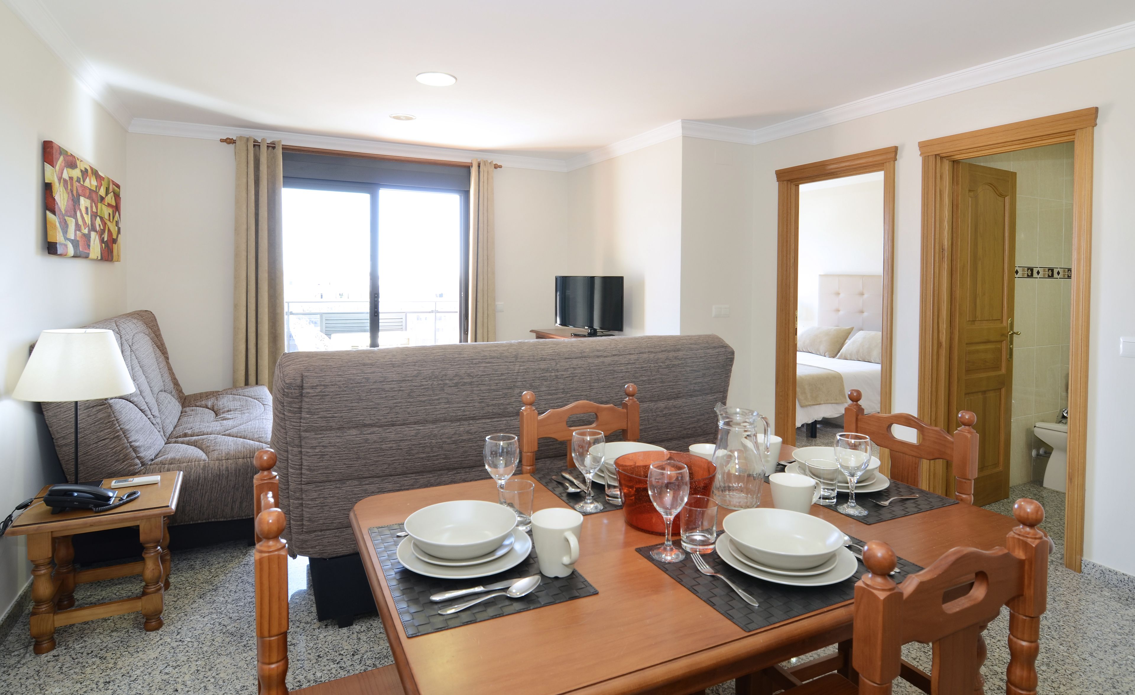 A hotel room with a dining area set for four, a sofa, a TV, and a bathroom visible through an open door.