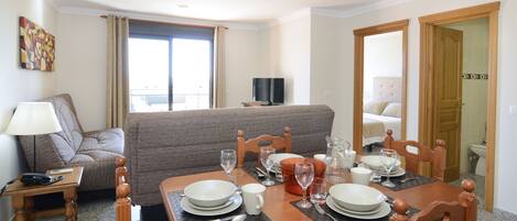A hotel room with a dining area set for four, a sofa, a TV, and a bathroom visible through an open door.