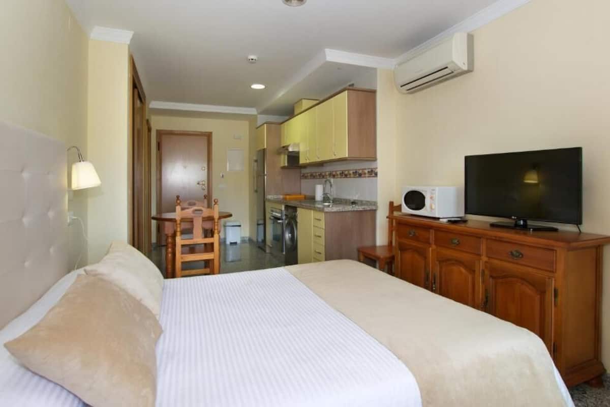 A hotel room with a bed, a television, a wooden dresser, and a kitchenette in the background.