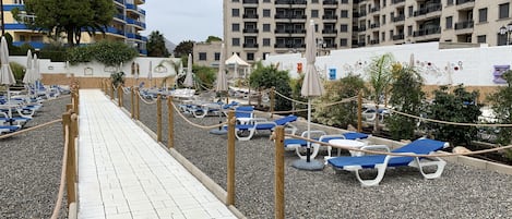 En sti ved stranden med liggestole og parasoller, omgivet af bygninger og grønt