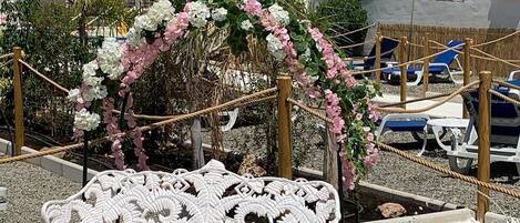 A white bench with intricate carvings, a rope barrier, and a decorative arch made of pink and white flowers.