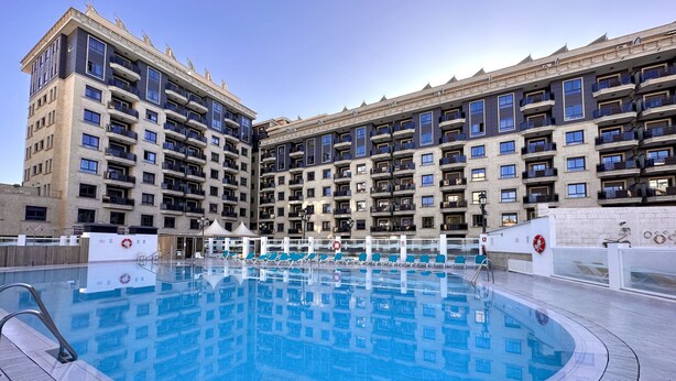 Un hotel moderno con una grande piscina esterna, numerosi balconi e cielo sereno.