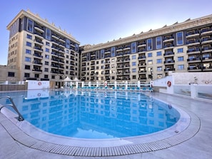 Un hotel moderno con una gran piscina al aire libre, varios balcones y un cielo despejado.