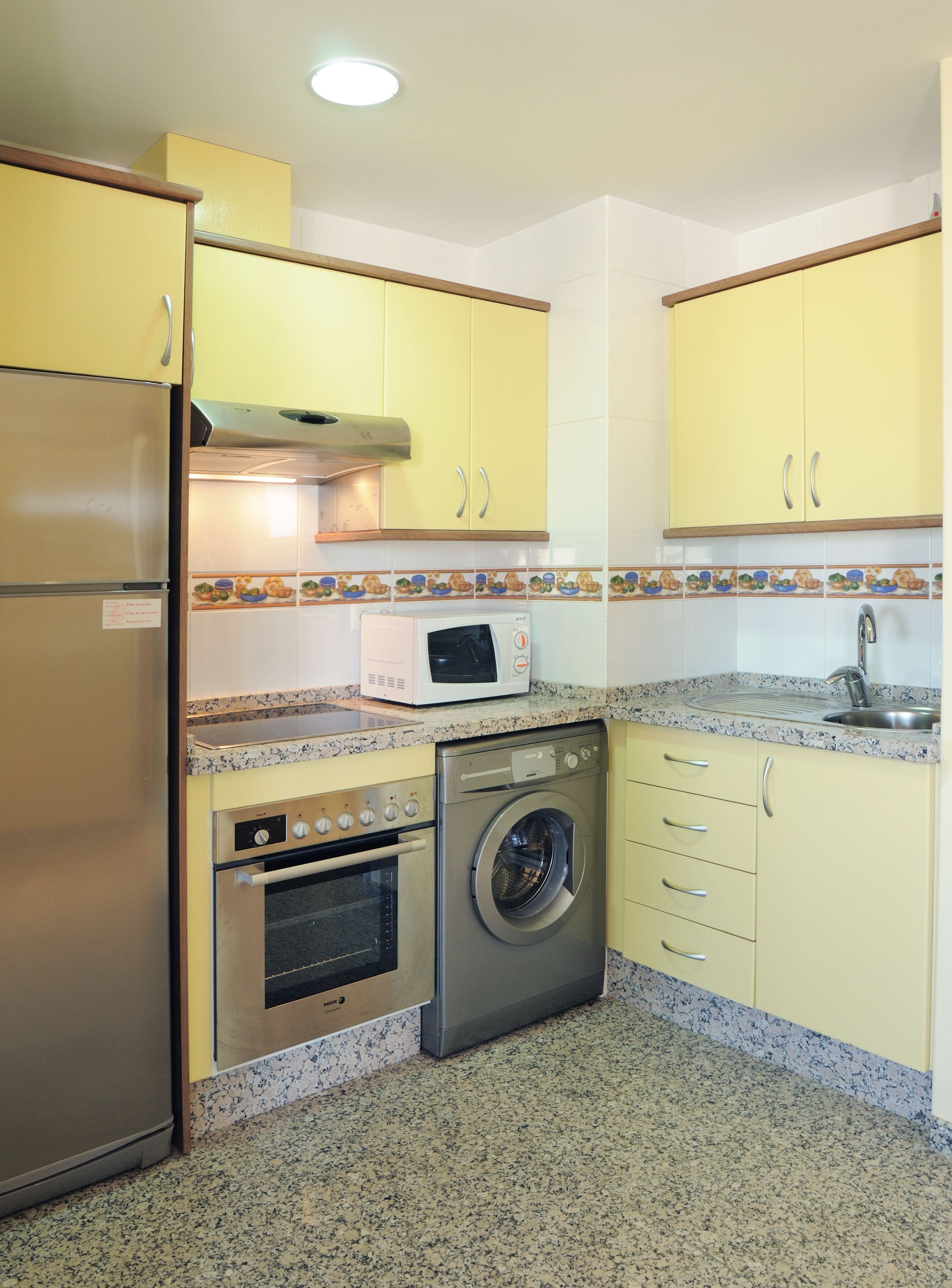 A compact kitchen with yellow cabinets, a stainless steel refrigerator, a microwave, an oven, and a washing machine.