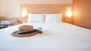 A hotel room with a large bed, two wall-mounted lights, a suitcase, and a woven hat on the bed. - ibis Porto Centro São Bento (Porto)
