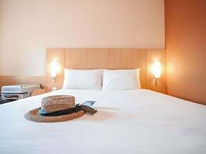 Quarto de hotel com uma cama grande, duas luminárias fixadas na parede, uma mala e um chapéu de palha sobre a cama.