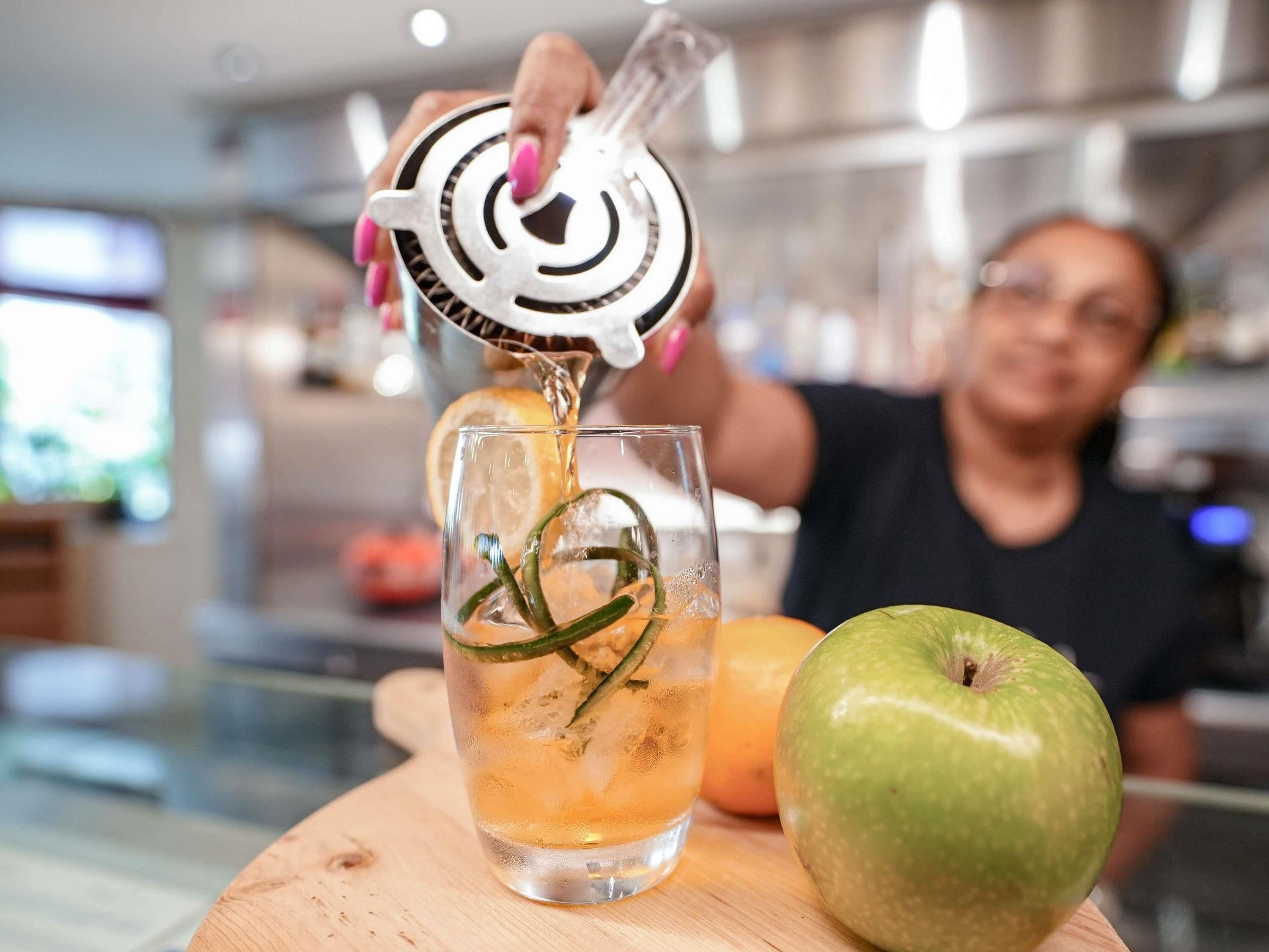 A person pouring a drink into a glass with a lemon slice and green garnish, with an apple and a wooden surface in the foreground.