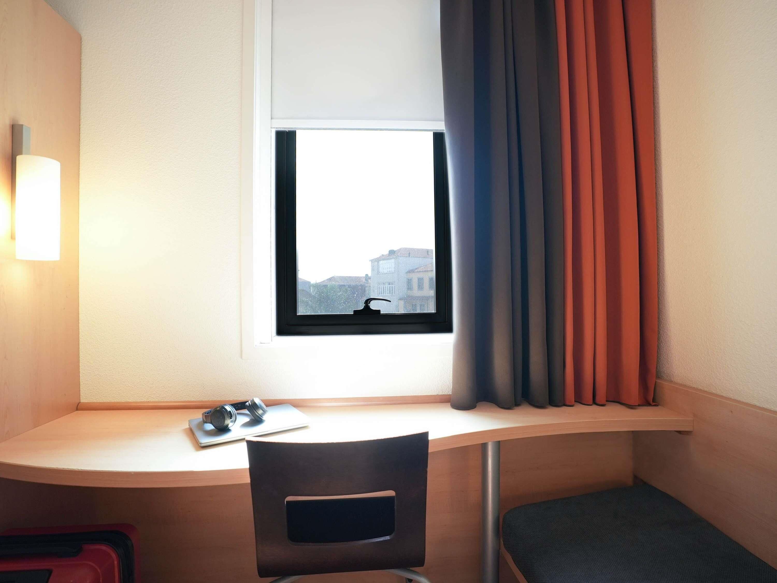 A hotel room with a desk, a chair, a window with curtains, and a wall-mounted lamp.