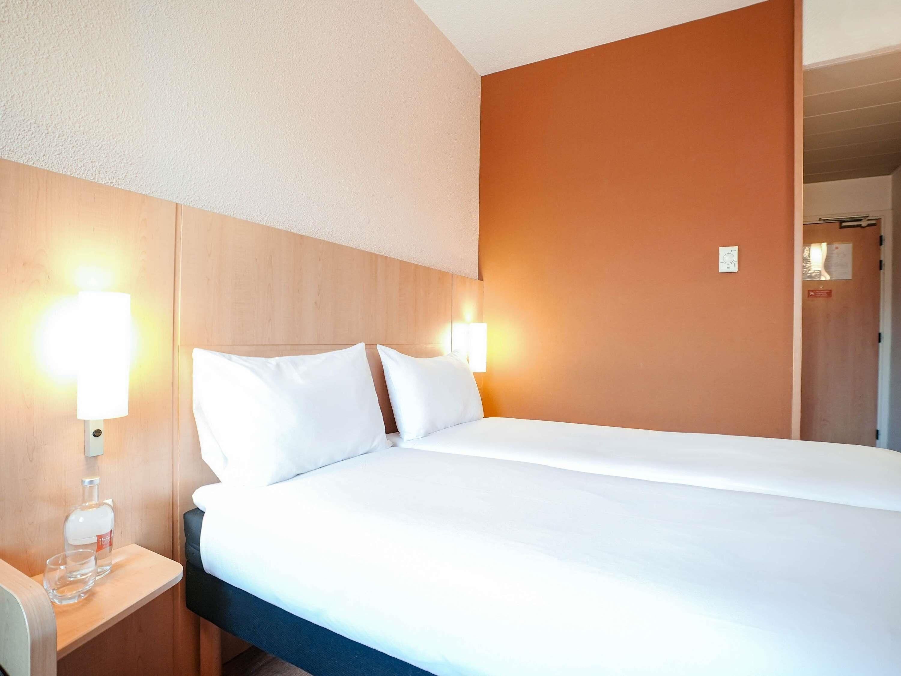 Hotellihuone, jossa on suuri sänky, kaksi yöpöytävalaisinta, yöpöytä, jossa on lasi vettä, ja puinen pääty.