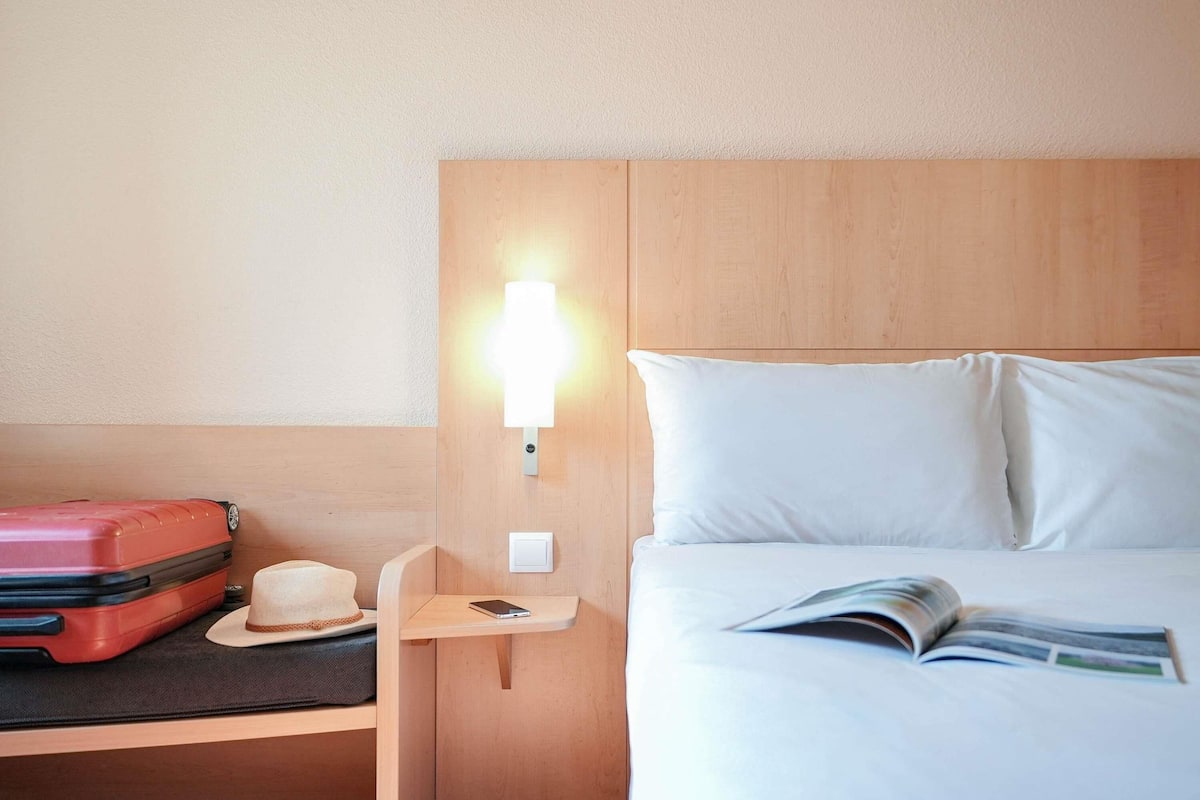 A hotel room with a bed, a nightstand, a red suitcase, a hat, and a magazine.