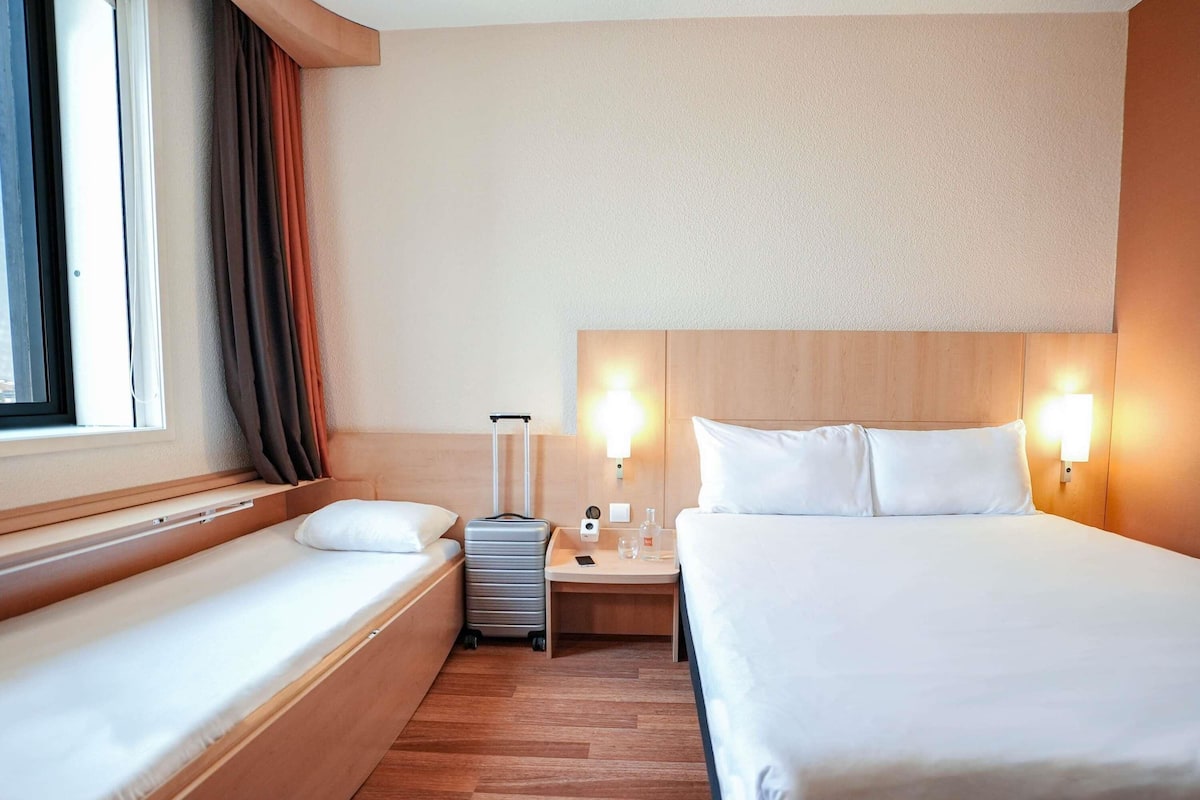 Ein Hotelzimmer mit Bett, kleinem Tisch mit Lampe, Nachttisch und einem Fenster mit Vorhängen.