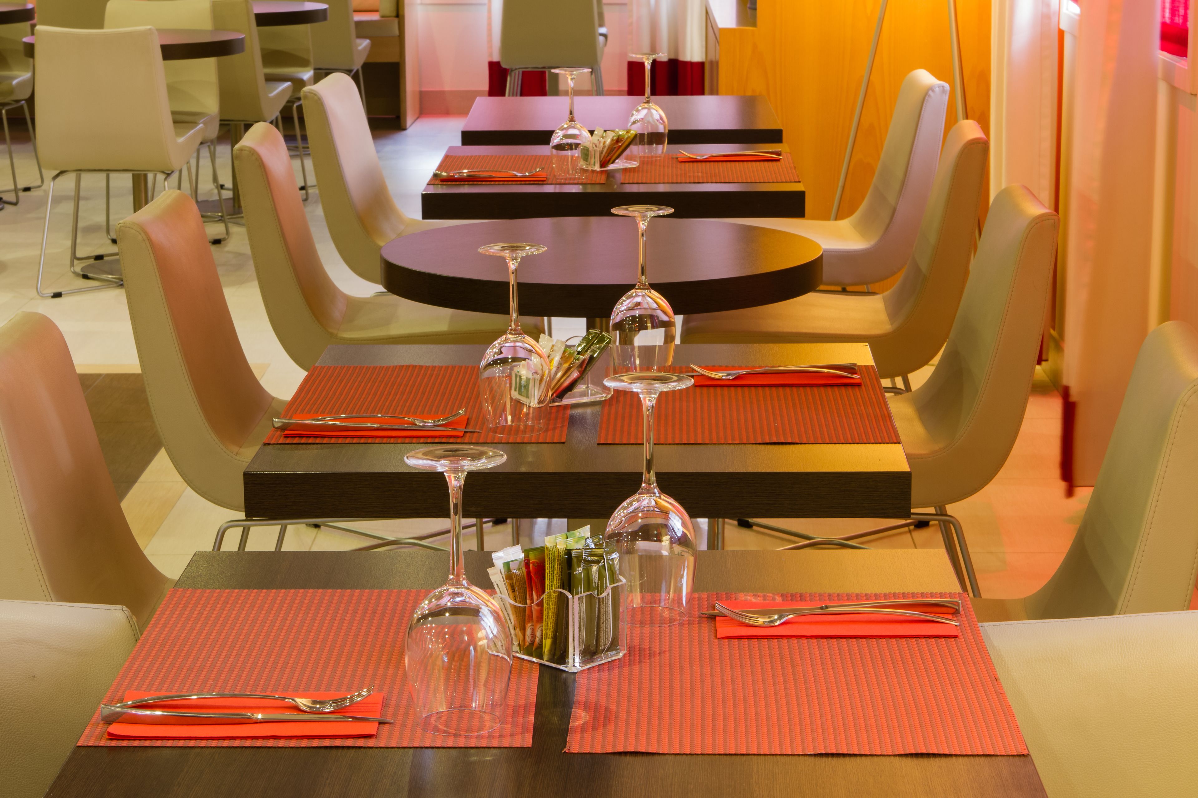 Un espace repas avec des tables préparées pour deux personnes, chacune agrémentée d’une paire de verres à vin, de couverts et de sets de table rouges.