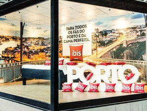 Quarto de hotel com uma janela grande com vista para a paisagem urbana, uma cama e vários travesseiros do hotel ibis no parapeito.