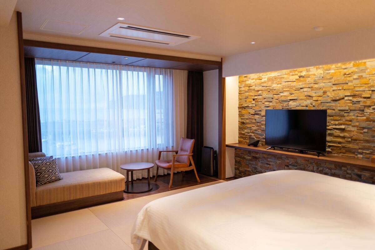 Une chambre d’hôtel comprenant un lit, un coin salon avec un canapé et un fauteuil, une petite table ronde, un téléviseur fixé à un mur en pierre et une grande fenêtre avec des rideaux.