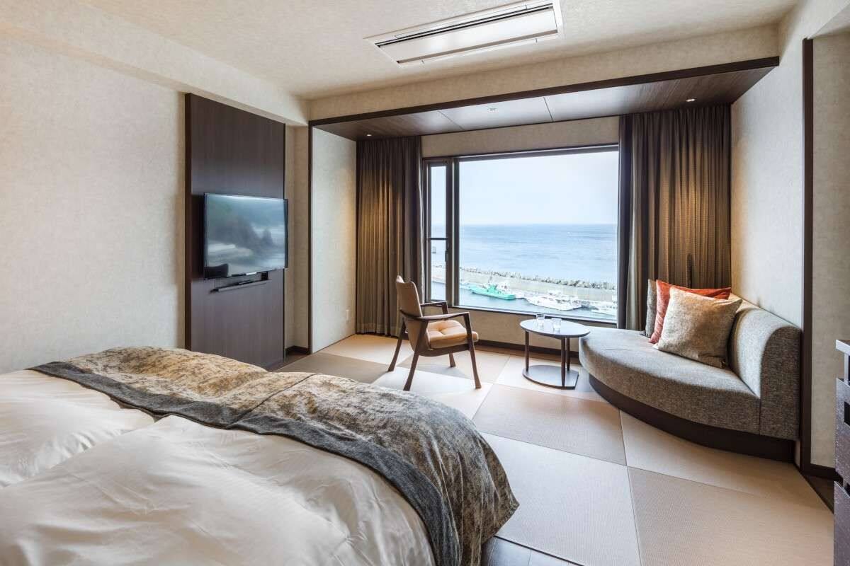 Une chambre d’hôtel avec un grand lit, un canapé, un fauteuil et une petite table. Il y a un téléviseur fixé au mur et une fenêtre donnant sur l’océan.