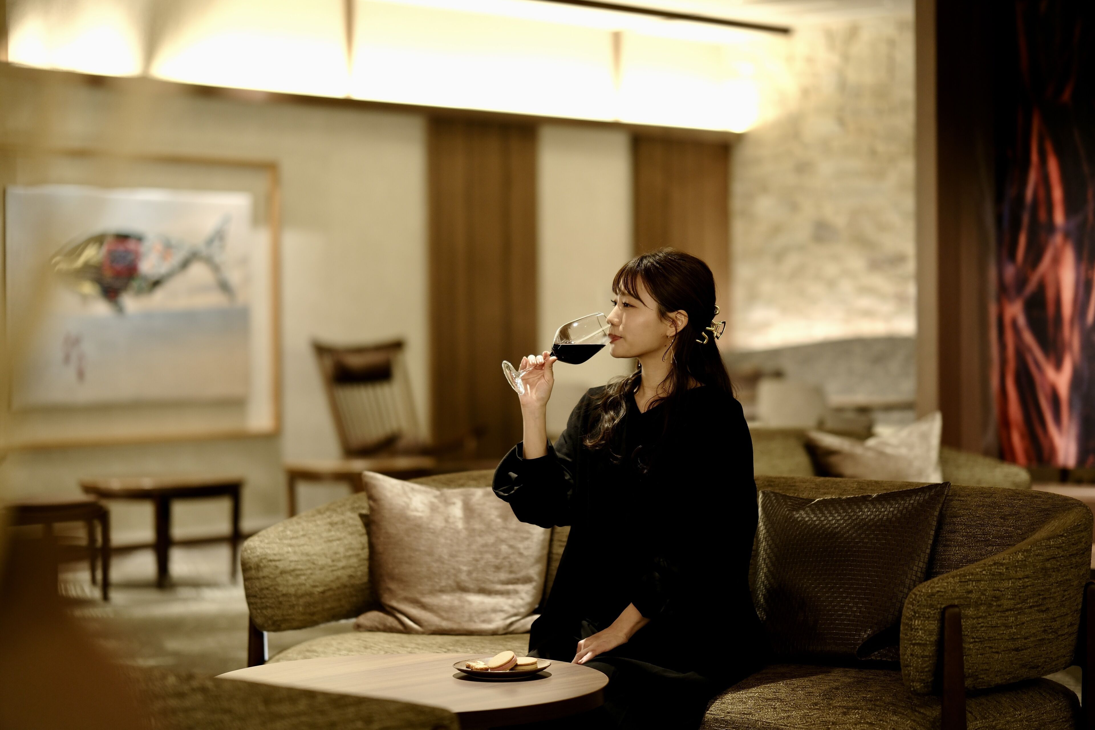 A woman sitting on a couch in a room with a table, a painting on the wall, and a lamp.